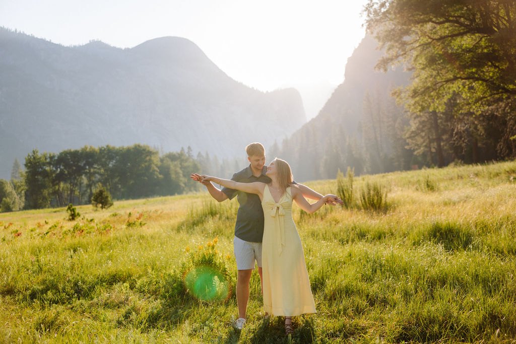 A woman in a yellow dress balances on a fallen tree while a man in shorts and a polo shirt holds her hand in a grassy valley with mountains in the background.