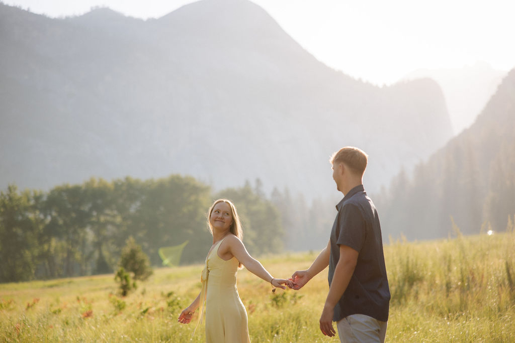 A couple stands outdoors, embracing with a scenic mountain and tree landscape in the background.