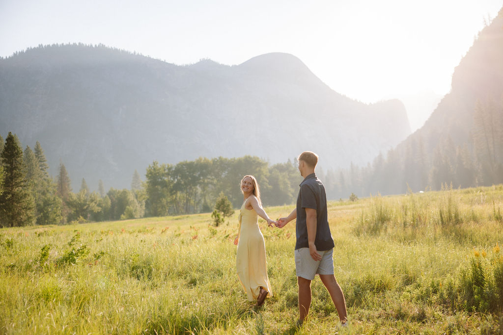 A couple stands outdoors, embracing with a scenic mountain and tree landscape in the background.