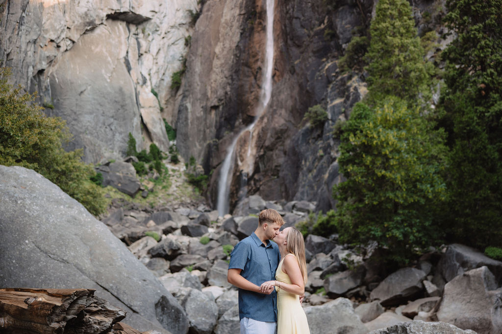 A couple stands embracing on rocks in front of a tall waterfall and cliffside, surrounded by trees and boulders.