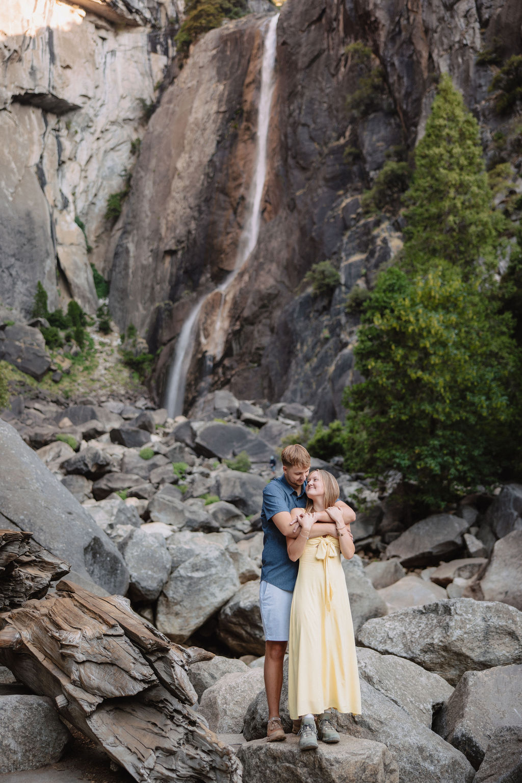 A couple stands embracing on rocks in front of a tall waterfall and cliffside, surrounded by trees and boulders.