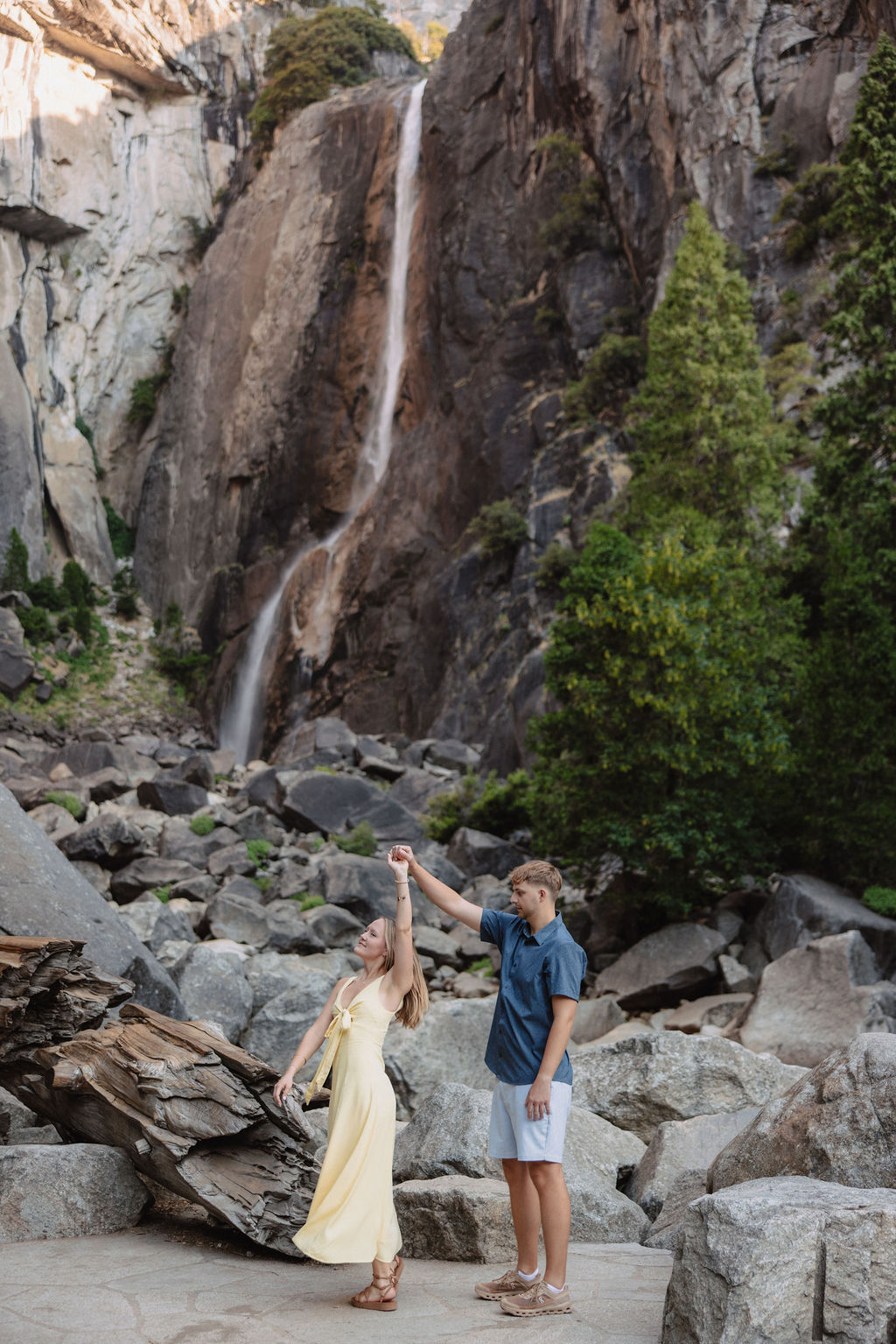 A couple stands embracing on rocks in front of a tall waterfall and cliffside, surrounded by trees and boulders.
