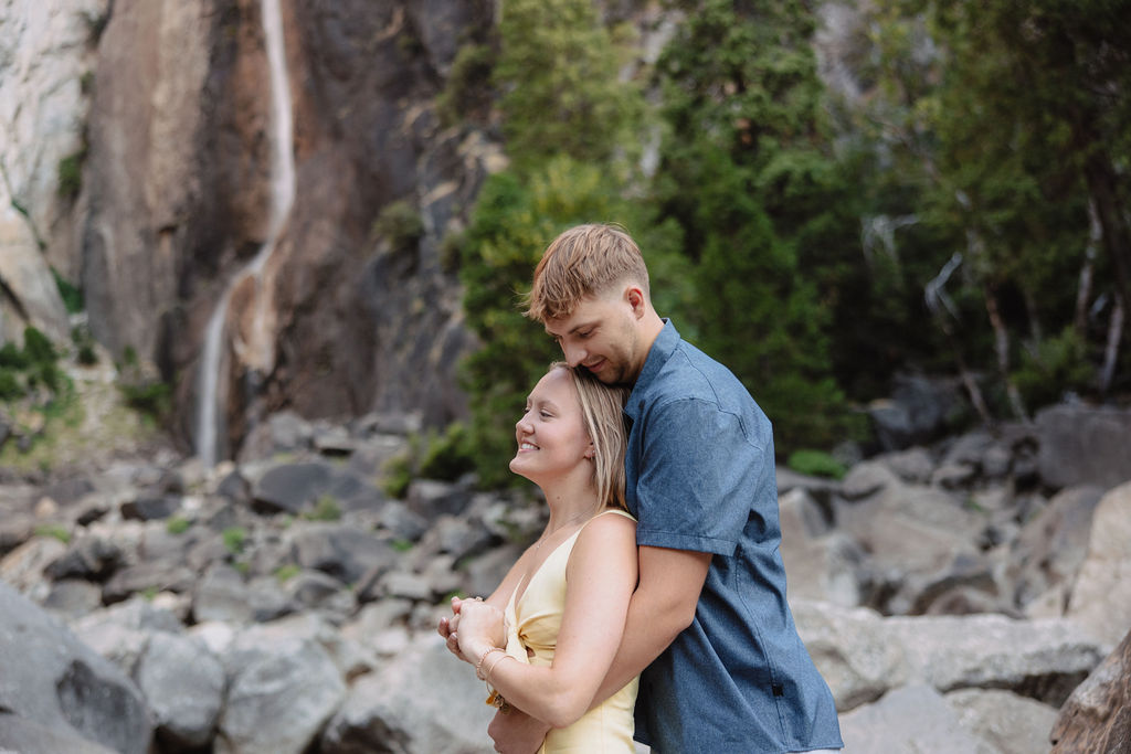 A couple stands embracing on rocks in front of a tall waterfall and cliffside, surrounded by trees and boulders.