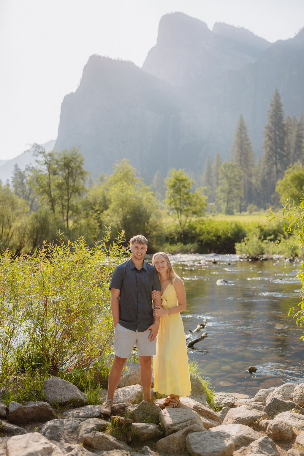 A man and a woman stand together by a river with trees and large mountains in the background on a sunny day.