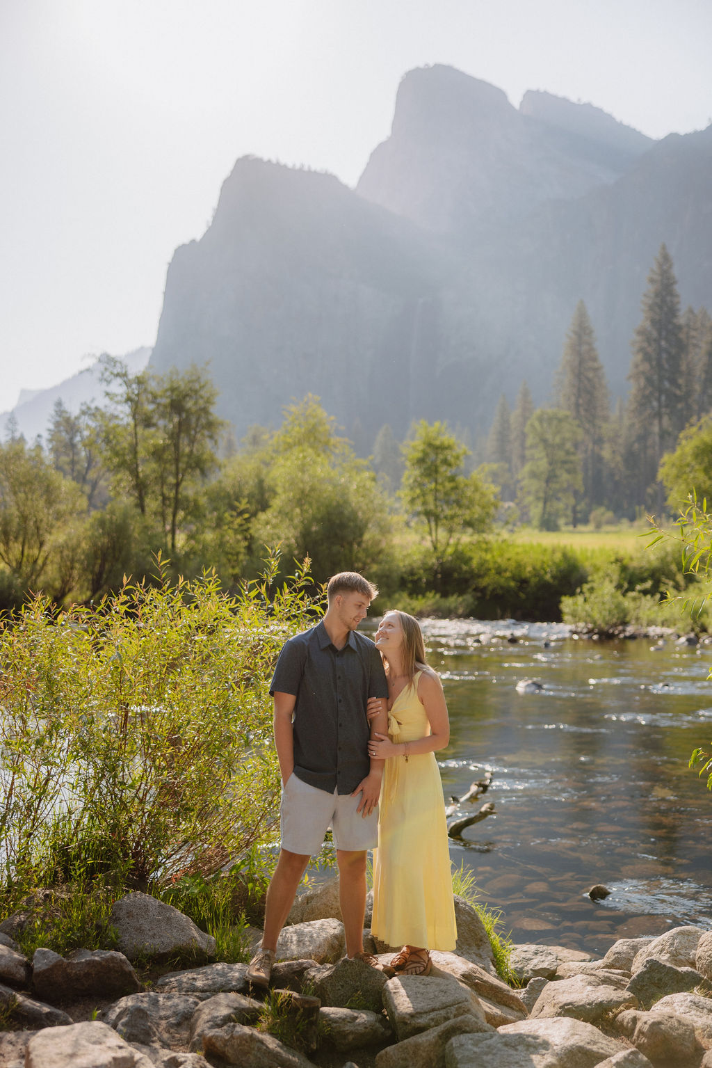 A man and a woman stand together by a river with trees and large mountains in the background on a sunny day.
