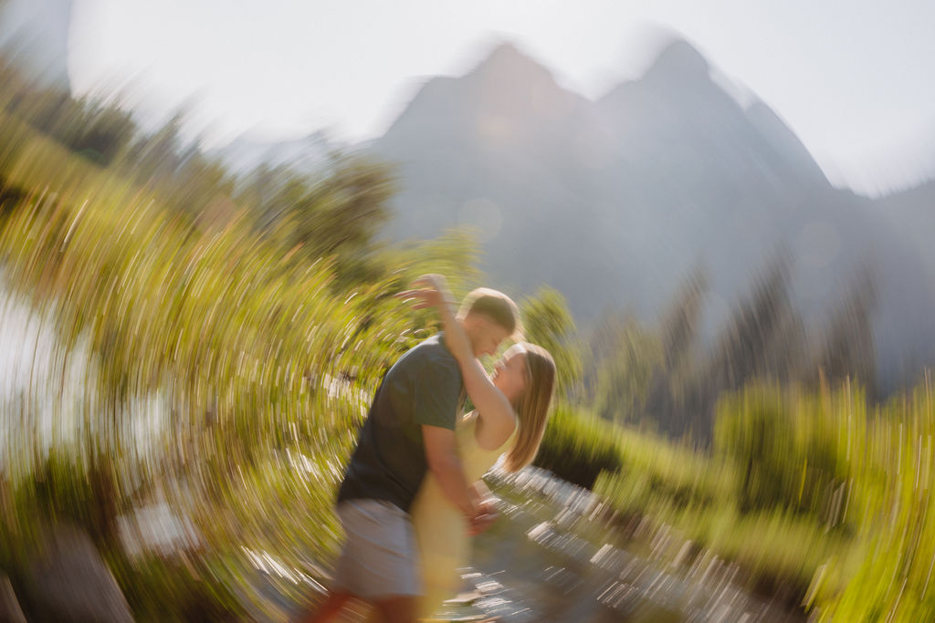A man and a woman stand together by a river with trees and large mountains in the background on a sunny day.