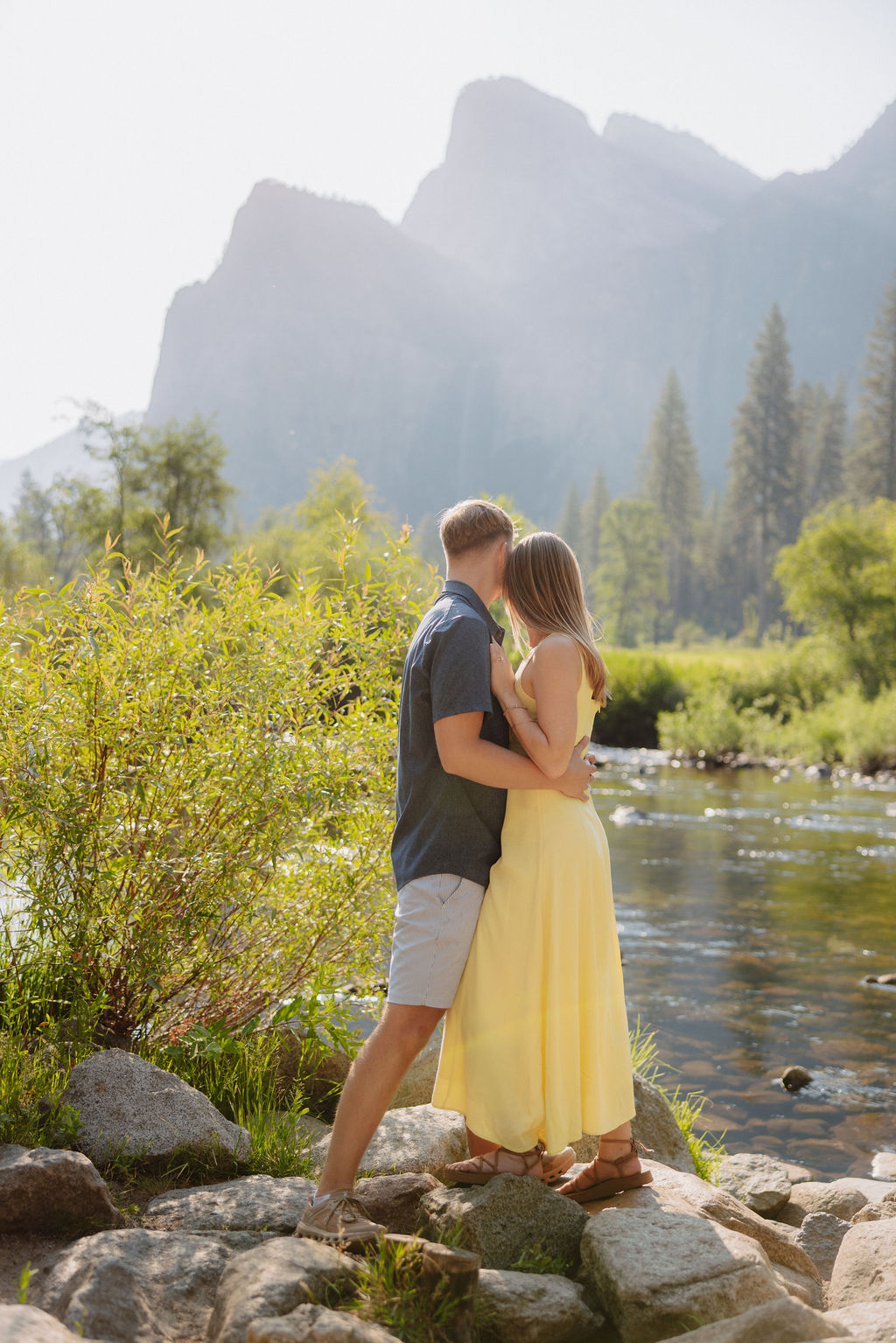 A man and a woman stand together by a river with trees and large mountains in the background on a sunny day.