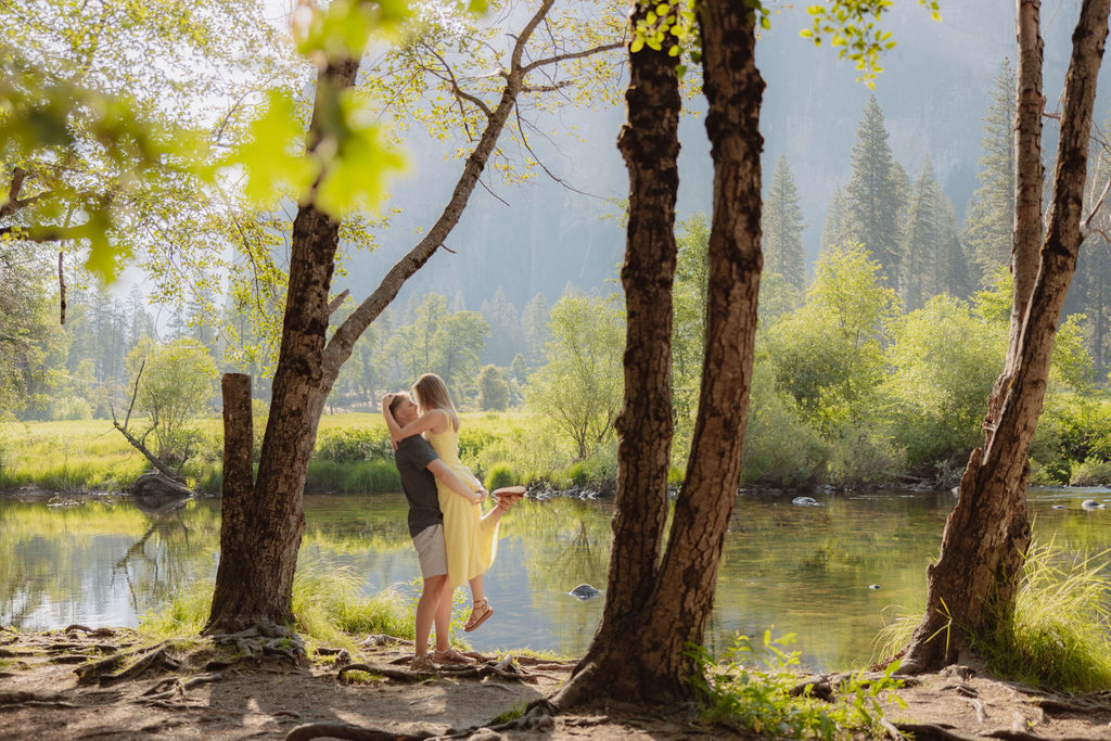 A man and a woman stand together by a river with trees and large mountains in the background on a sunny day.
