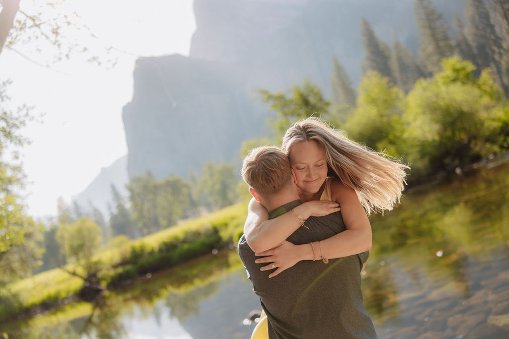 A man and a woman stand together by a river with trees and large mountains in the background on a sunny day.