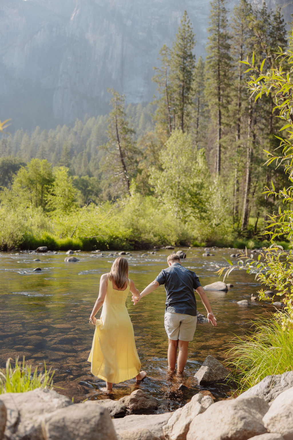 A man and a woman stand together by a river with trees and large mountains in the background on a sunny day.