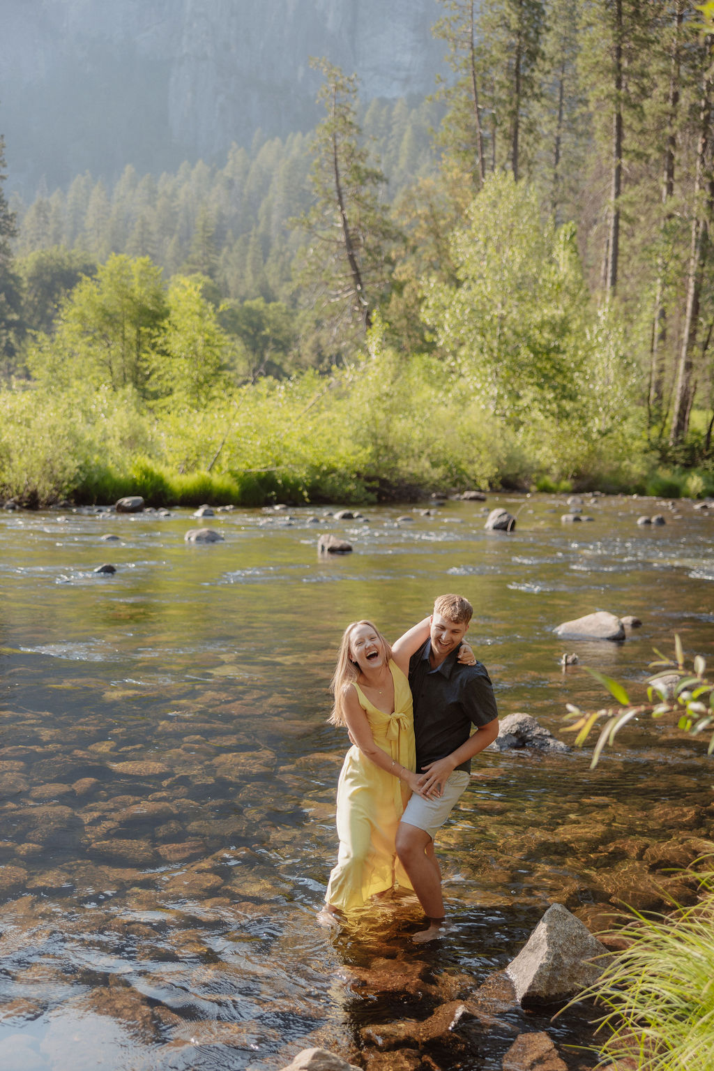 A man and a woman stand together by a river with trees and large mountains in the background on a sunny day.
