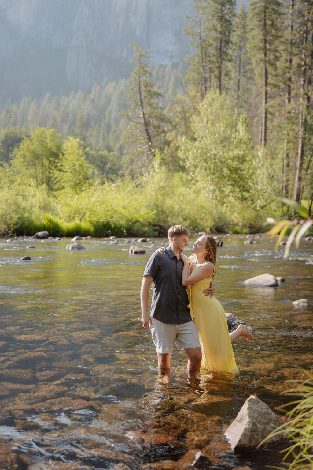 A man and a woman stand together by a river with trees and large mountains in the background on a sunny day.