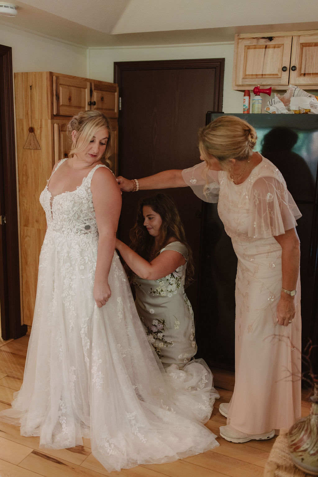 A bride in a white gown is being assisted by two women in light-colored dresses as she gets ready indoors.