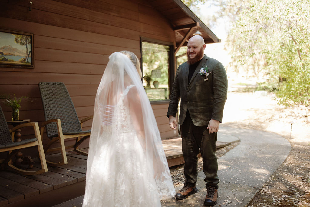 A bride and groom stand facing each other outside a small wooden cabin surrounded by trees.