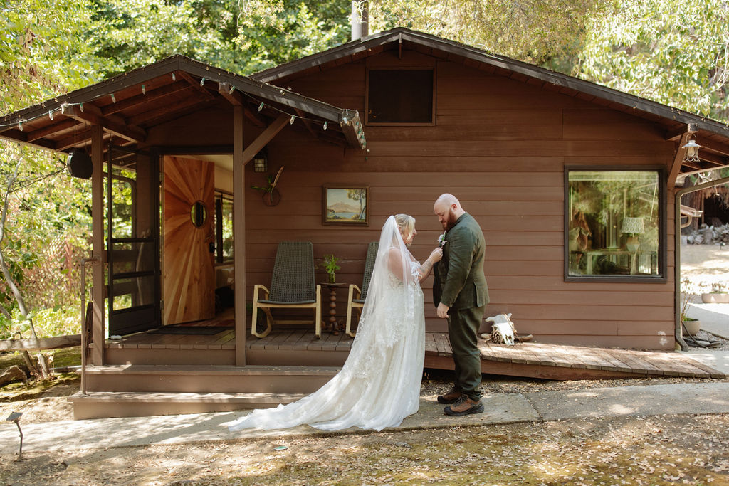 A bride and groom stand facing each other outside a small wooden cabin surrounded by trees.