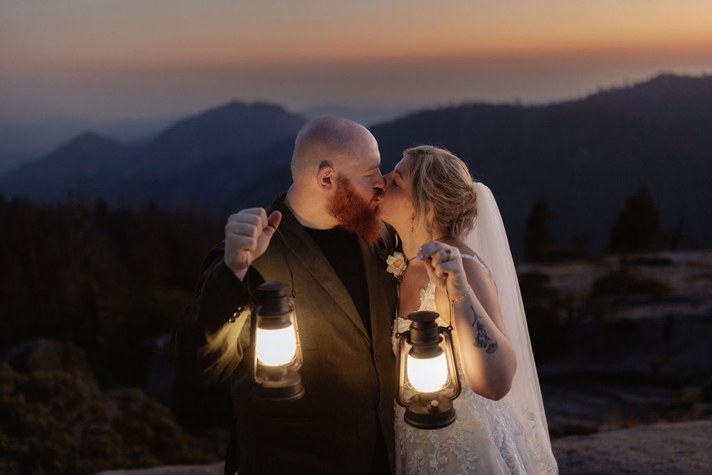 A bride and groom stand on a rocky ledge at sunset, facing the mountains and holding lanterns, with the bride’s veil flowing behind her.