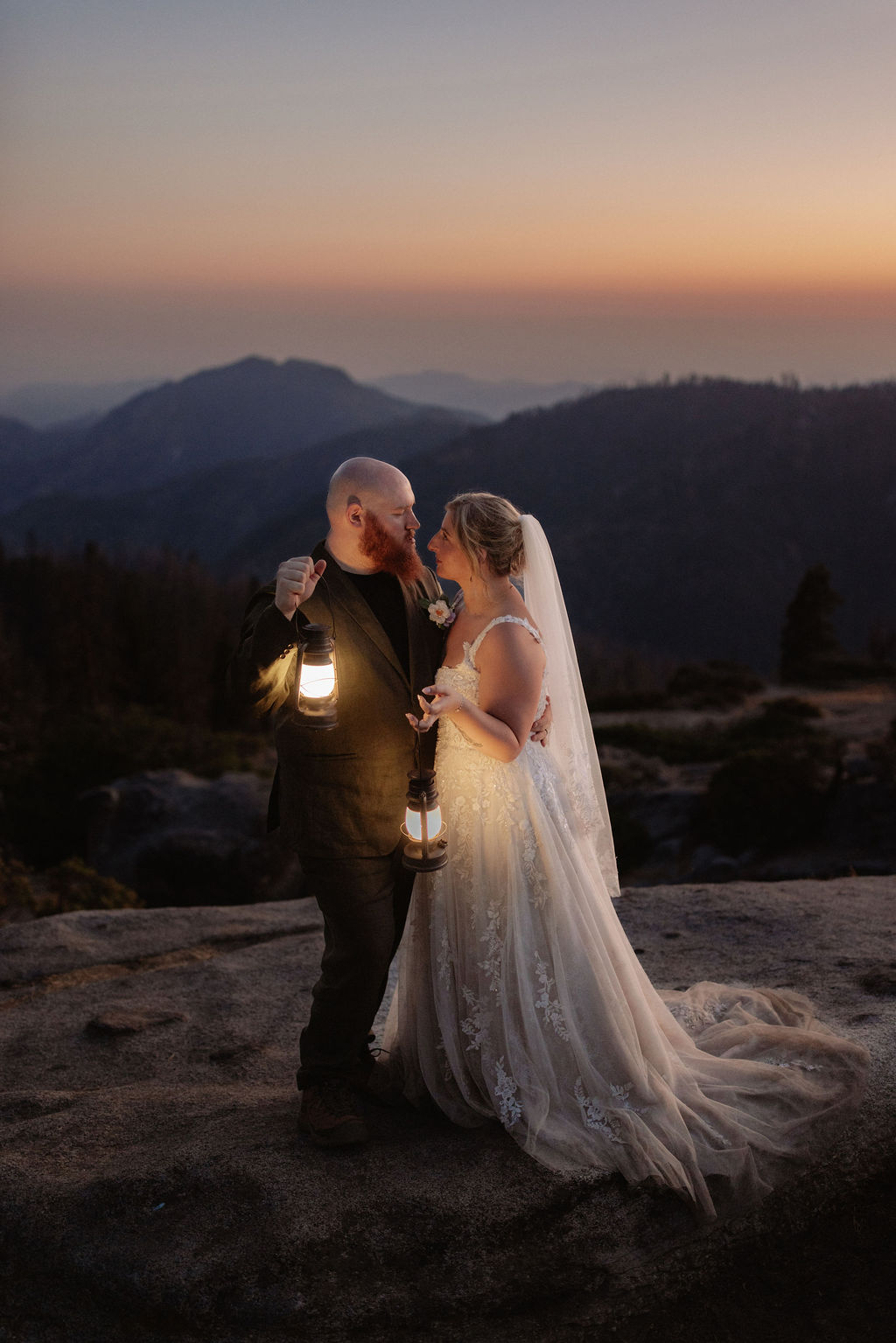 A bride and groom stand on a rocky ledge at sunset, facing the mountains and holding lanterns, with the bride’s veil flowing behind her.