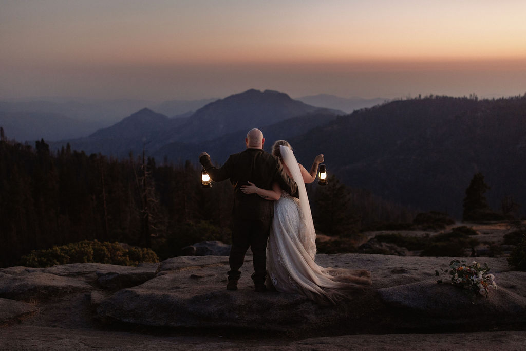 A bride and groom stand on a rocky ledge at sunset, facing the mountains and holding lanterns, with the bride’s veil flowing behind her.