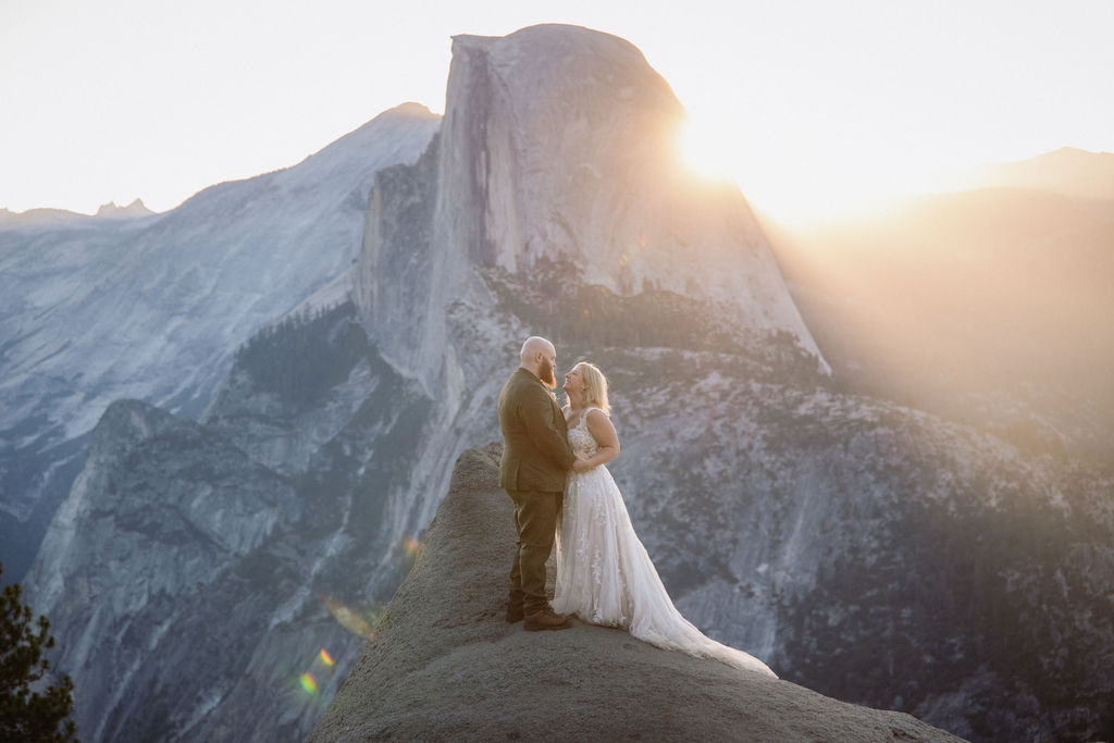 A couple in wedding attire stands on a rocky ledge with mountains and sunlight in the background for an elopement in Yosemite and Sequoia