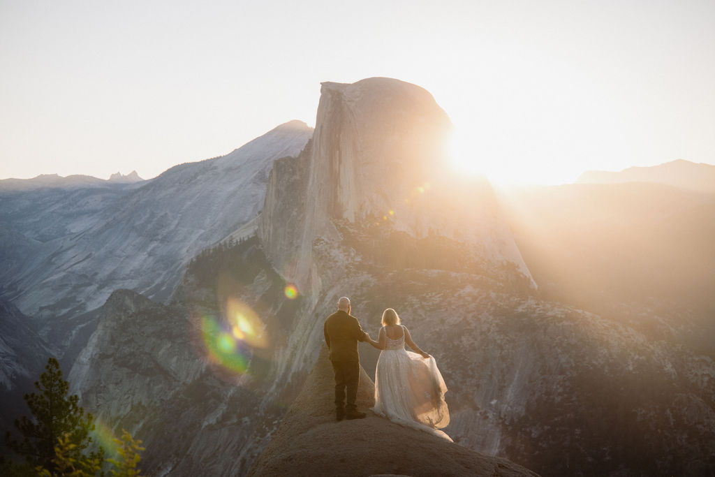 A couple in wedding attire stands on a rocky ledge with mountains and sunlight in the background for an elopement in Yosemite and Sequoia