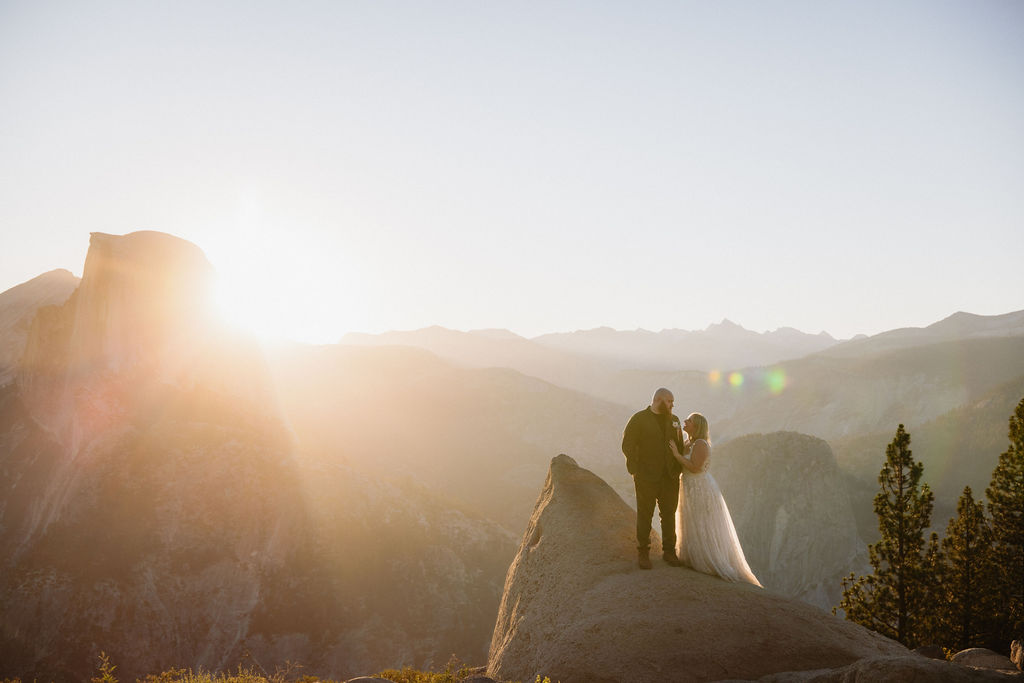 A couple in wedding attire stands on a rocky ledge with mountains and sunlight in the background for an elopement in Yosemite and Sequoia