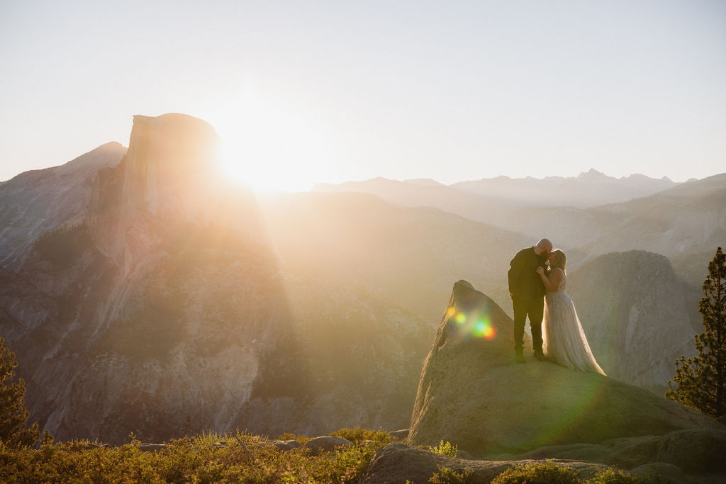 A couple in wedding attire stands on a rocky ledge with mountains and sunlight in the background for an elopement in Yosemite and Sequoia