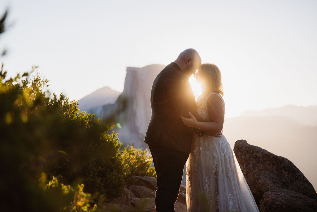 A couple in wedding attire stands on a rocky ledge with mountains and sunlight in the background for an elopement in Yosemite and Sequoia