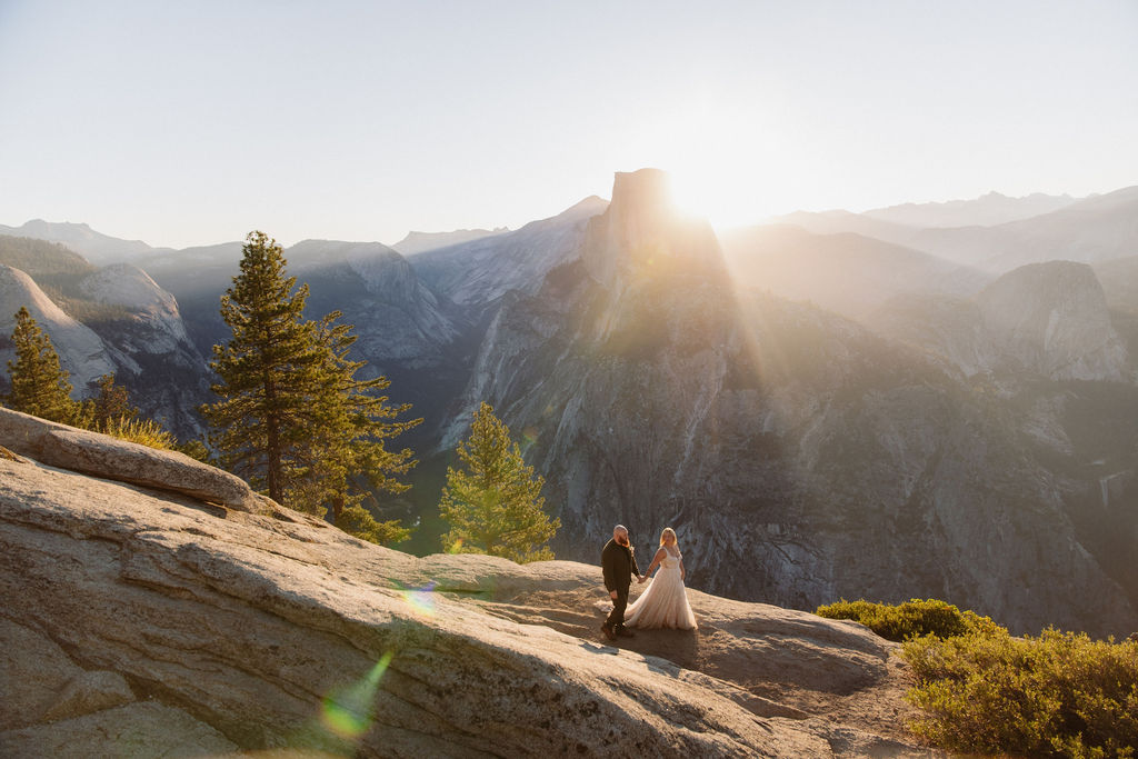 A couple in wedding attire stands on a rocky ledge with mountains and sunlight in the background for an elopement in Yosemite and Sequoia