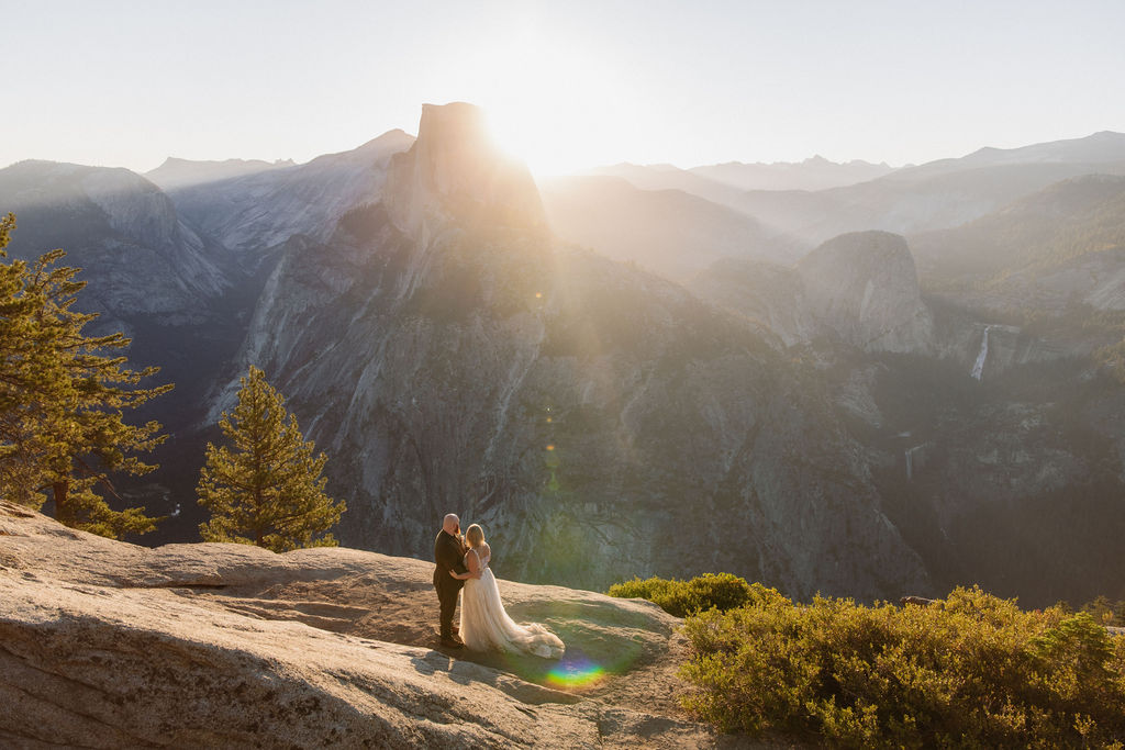 A couple in wedding attire stands on a rocky ledge with mountains and sunlight in the background for an elopement in Yosemite and Sequoia