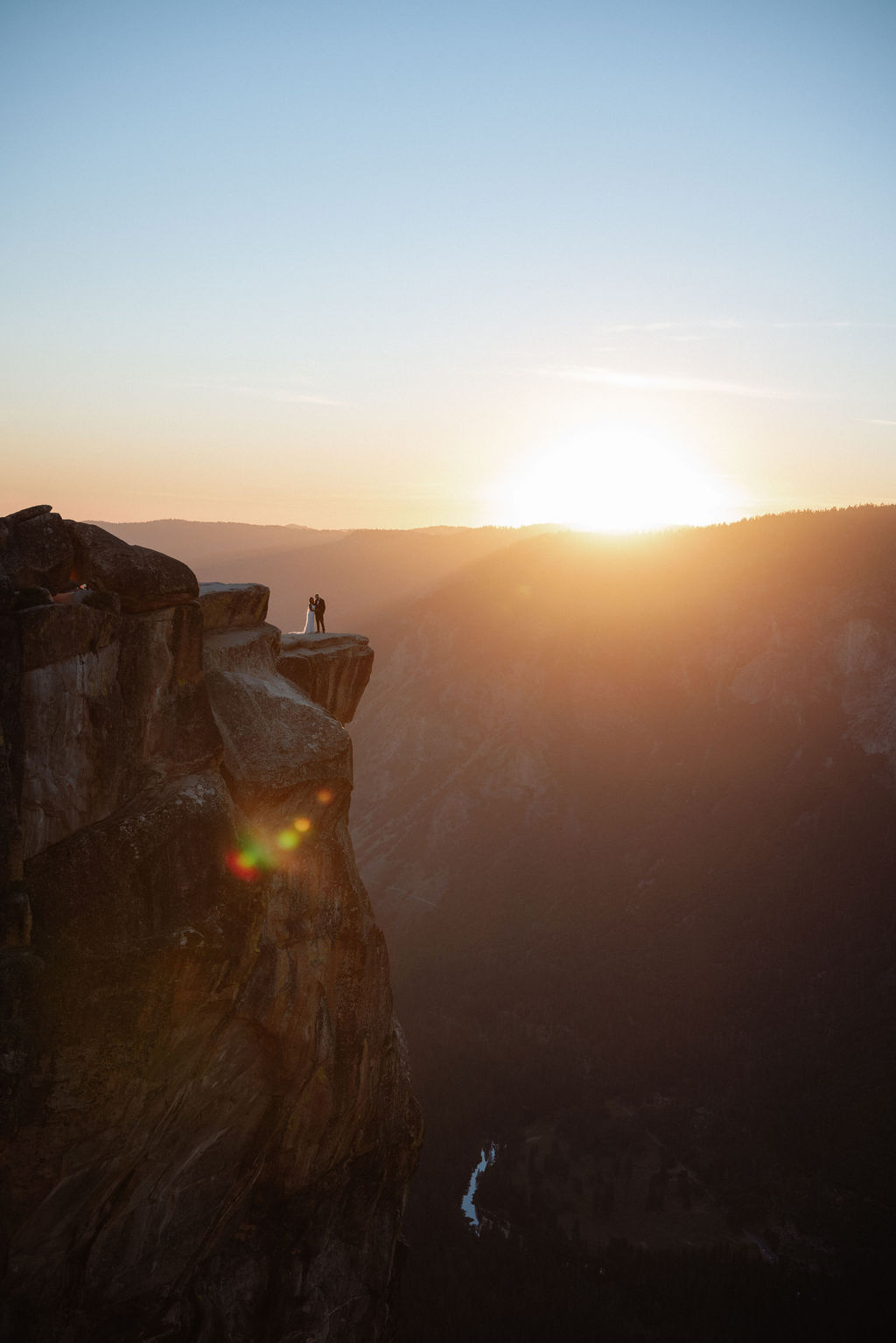 A person stands on the edge of a rocky cliff at sunset, overlooking a vast valley with distant mountains and a river below for their unique elopement