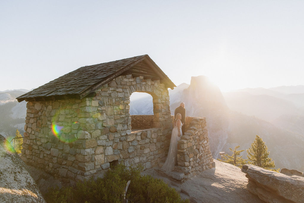 A stone lookout structure sits on a rocky ledge at sunrise, with a couple standing by the wall and mountains visible in the background.