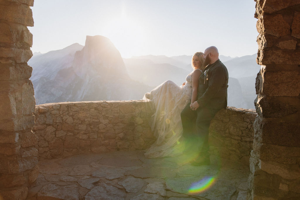 A stone lookout structure sits on a rocky ledge at sunrise, with a couple standing by the wall and mountains visible in the background.