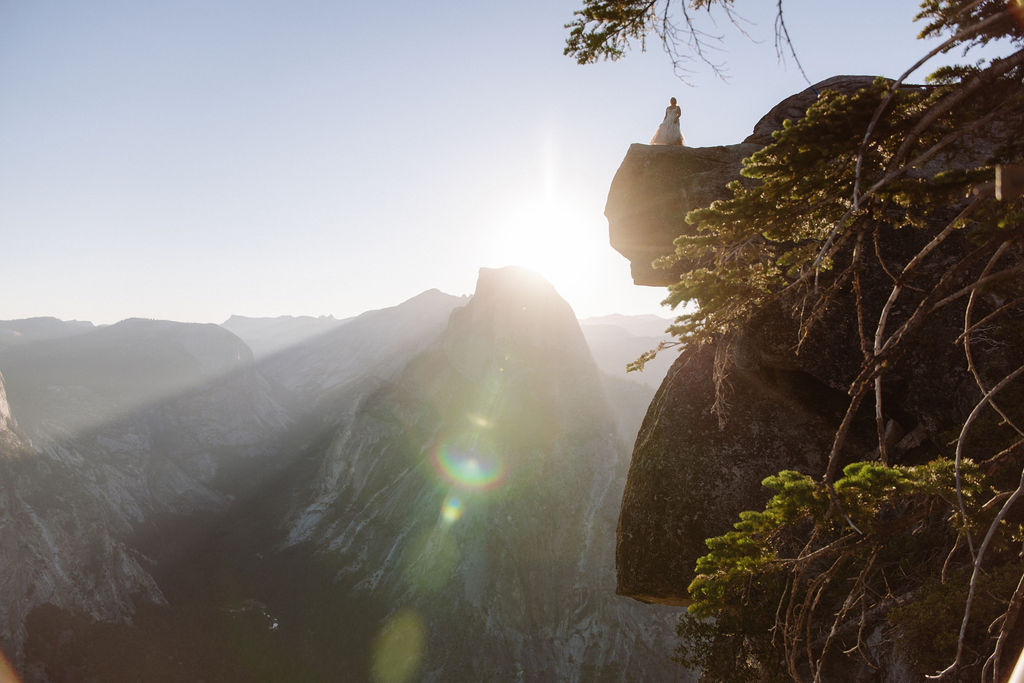Person sitting on a rocky cliff edge overlooking a valley with mountains in the background, sun shining low and bright in the sky.
