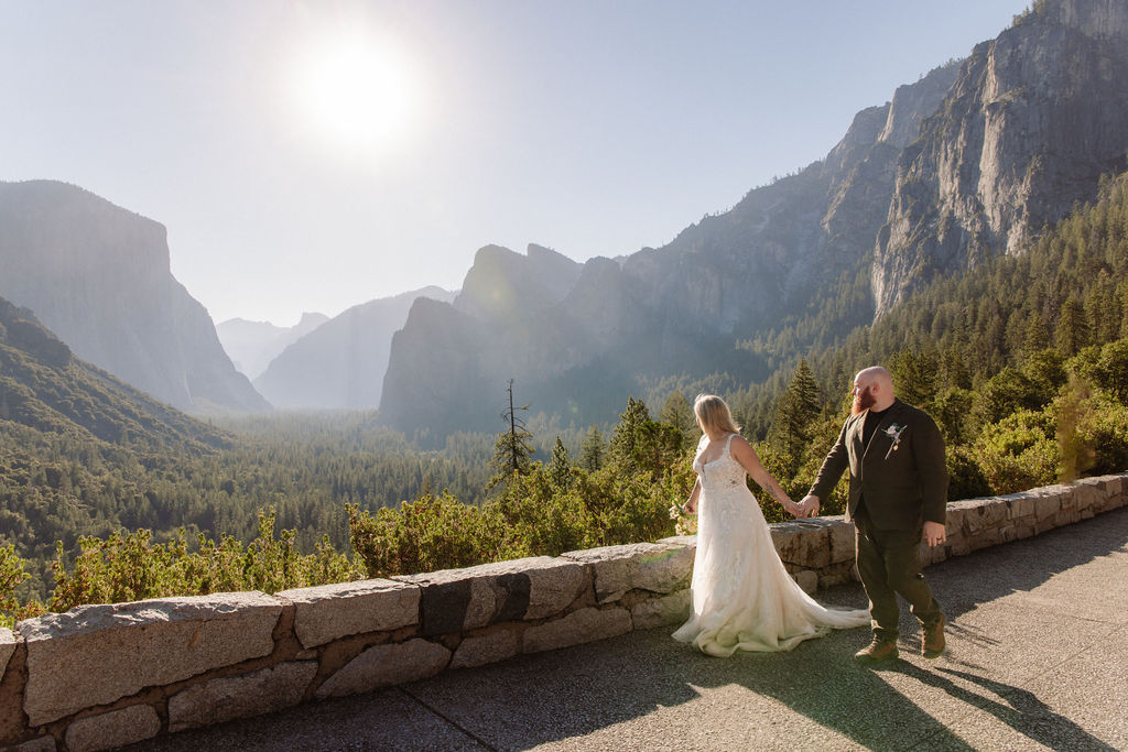 A bride and groom stand together outdoors surrounded by trees with a large rock formation and river in the background, under a bright sky.