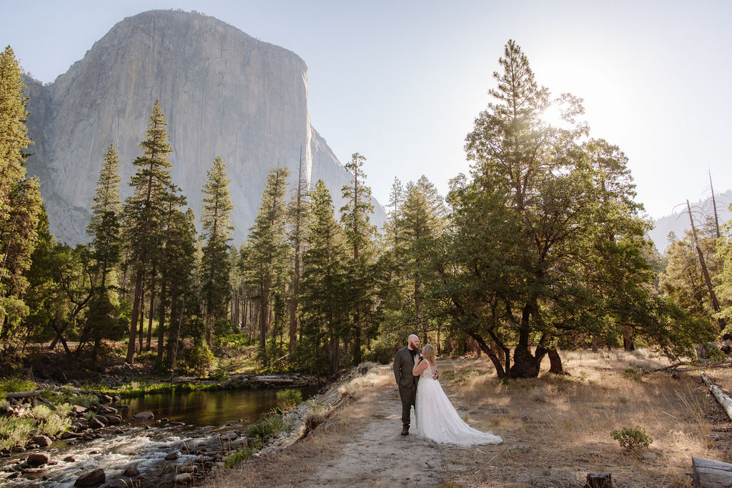 A bride and groom stand together outdoors surrounded by trees with a large rock formation and river in the background, under a bright sky.