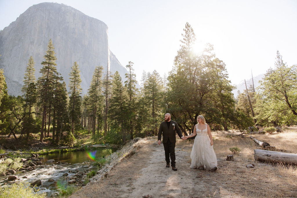 A bride and groom stand together outdoors surrounded by trees with a large rock formation and river in the background, under a bright sky.