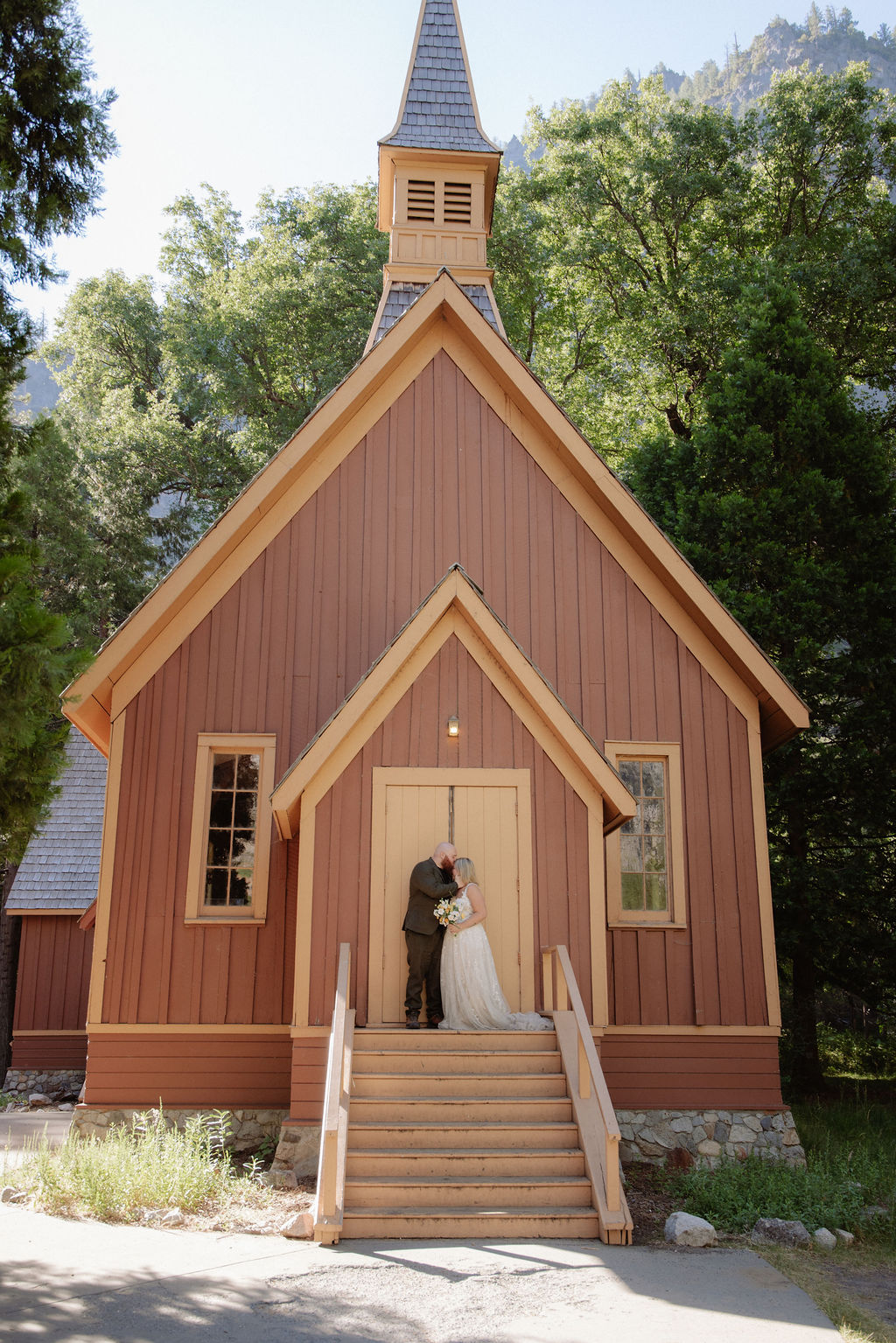 A bride and groom stand on the steps outside a small brown wooden chapel surrounded by trees.