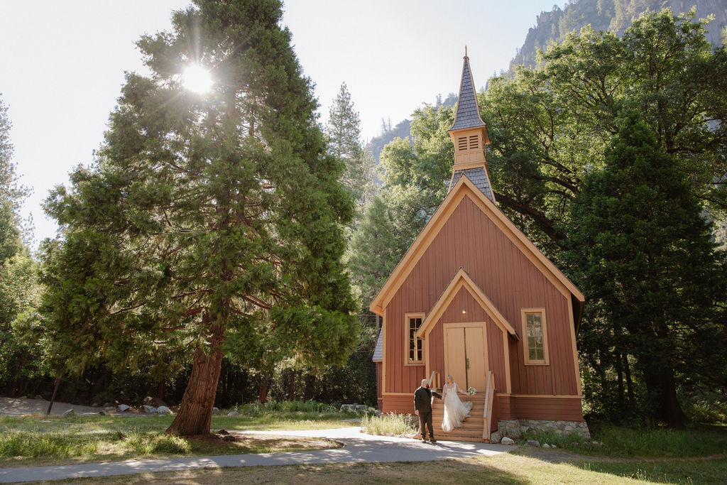 A bride and groom stand on the steps outside a small brown wooden chapel surrounded by trees.