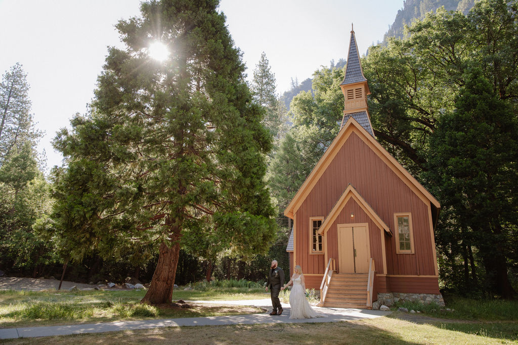 A bride and groom stand on the steps outside a small brown wooden chapel surrounded by trees.