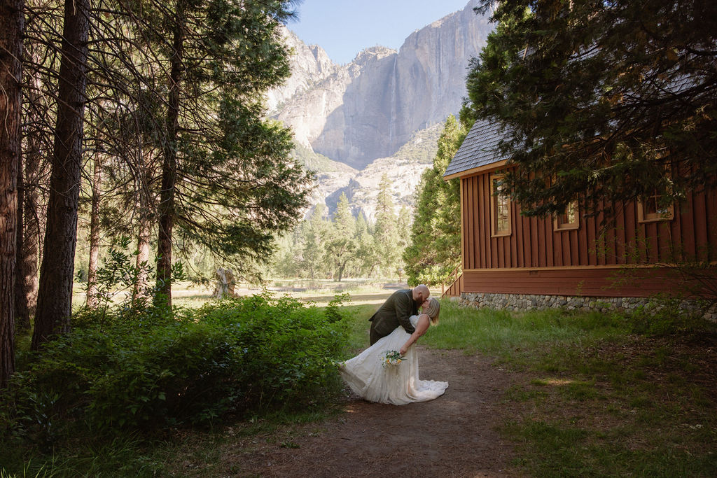 A bride and groom share a kiss outdoors near a wooden building, surrounded by tall trees and mountains in the background.