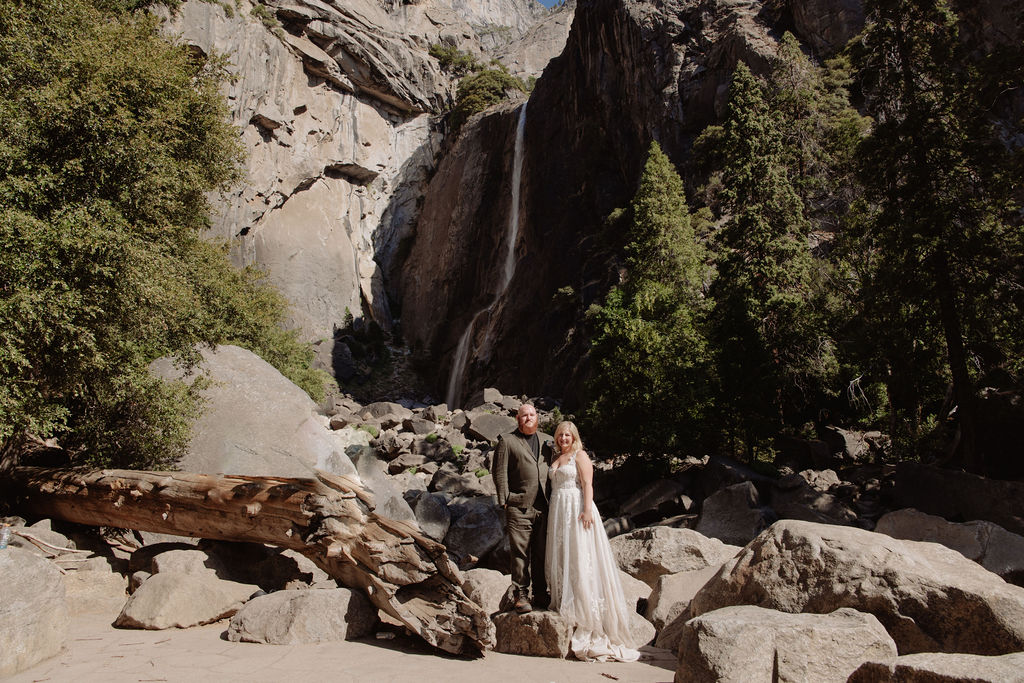 A bride and groom stand together on rocks in front of a waterfall and large trees in a mountainous outdoor setting.