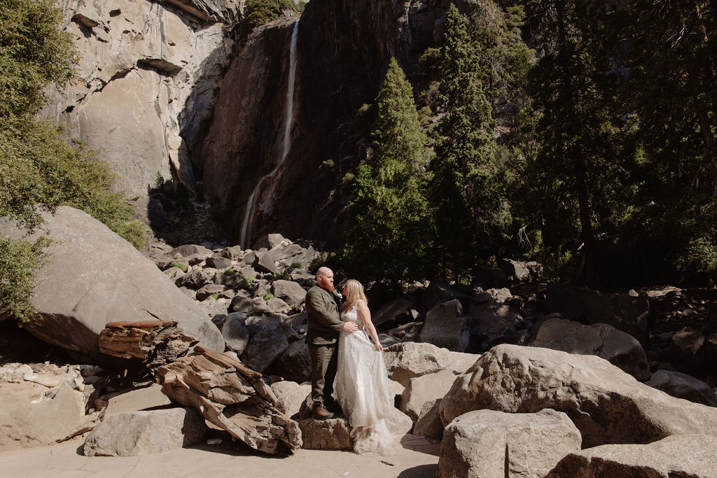 A bride and groom stand together on rocks in front of a waterfall and large trees in a mountainous outdoor setting.