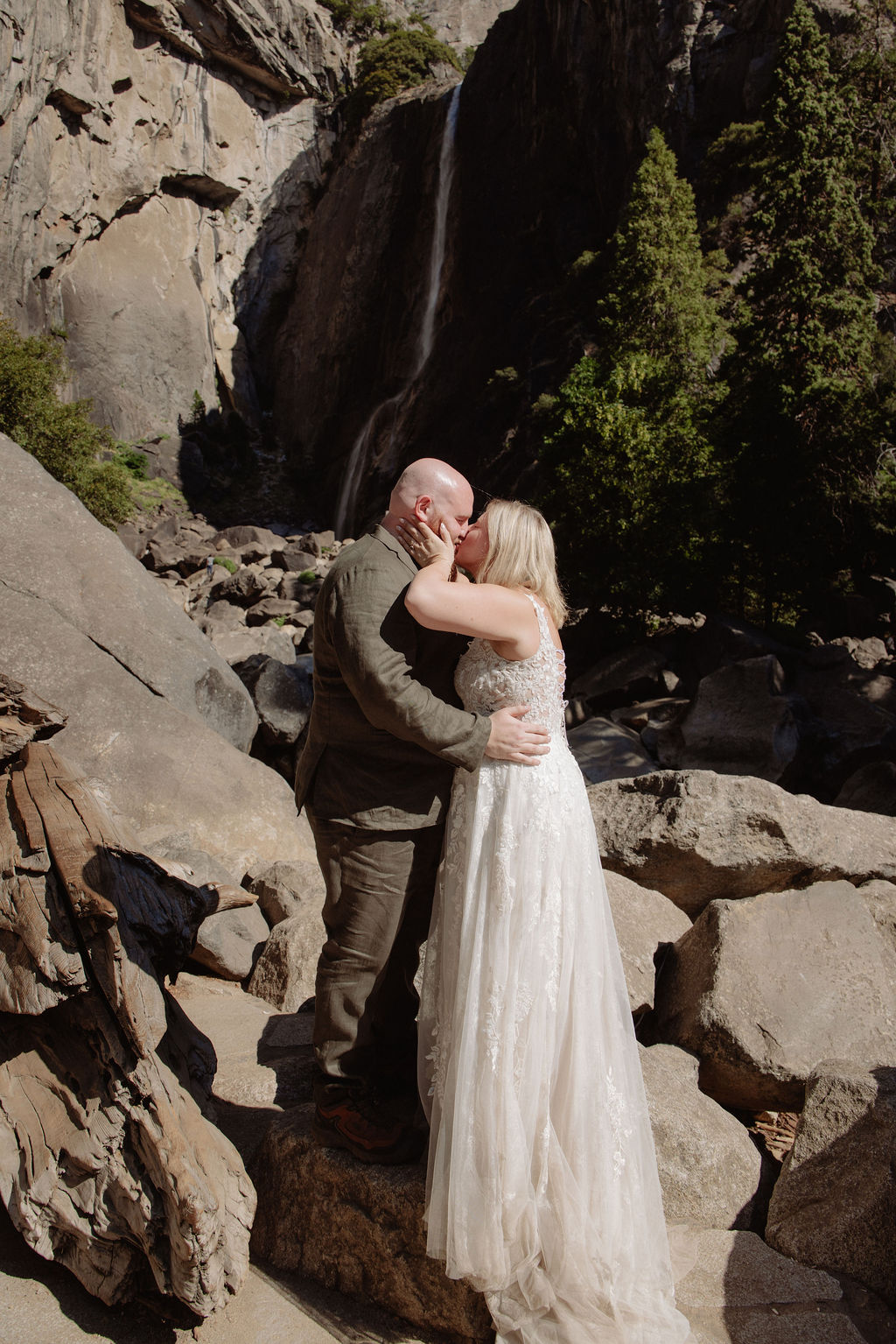 A bride and groom stand together on rocks in front of a waterfall and large trees in a mountainous outdoor setting.