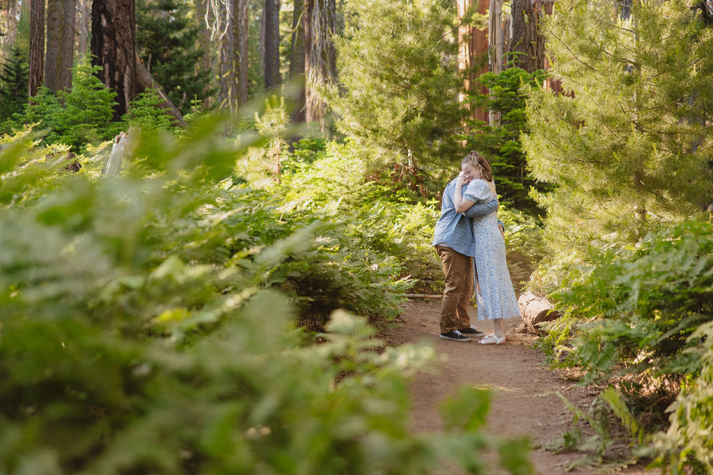 A man and woman walk hand in hand on a dirt path through a forest with tall trees and lush greenery.