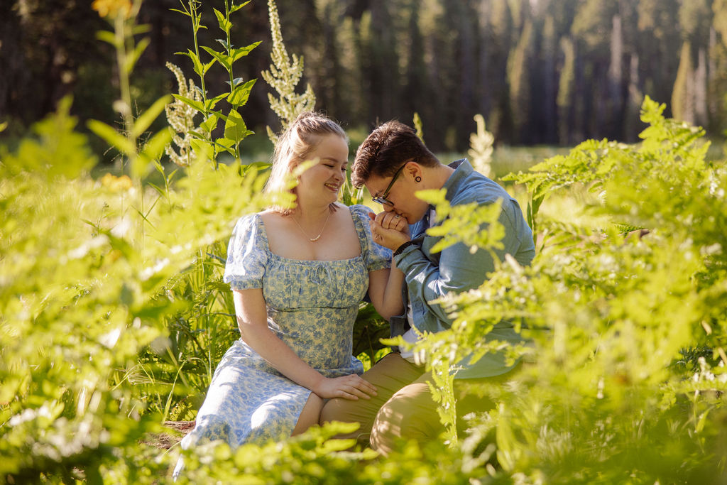 Two people stand in a sunlit forest clearing; one is helping the other step down from a log as they hold hands among tall green plants.