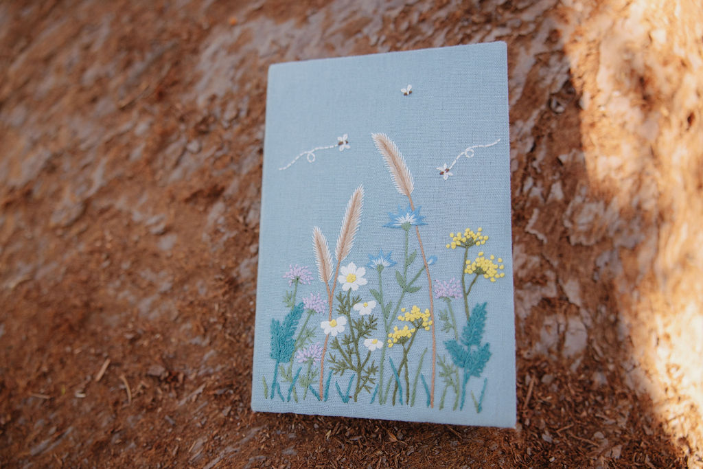 A blue canvas featuring an embroidered design of wildflowers and grasses rests against a textured brown surface.