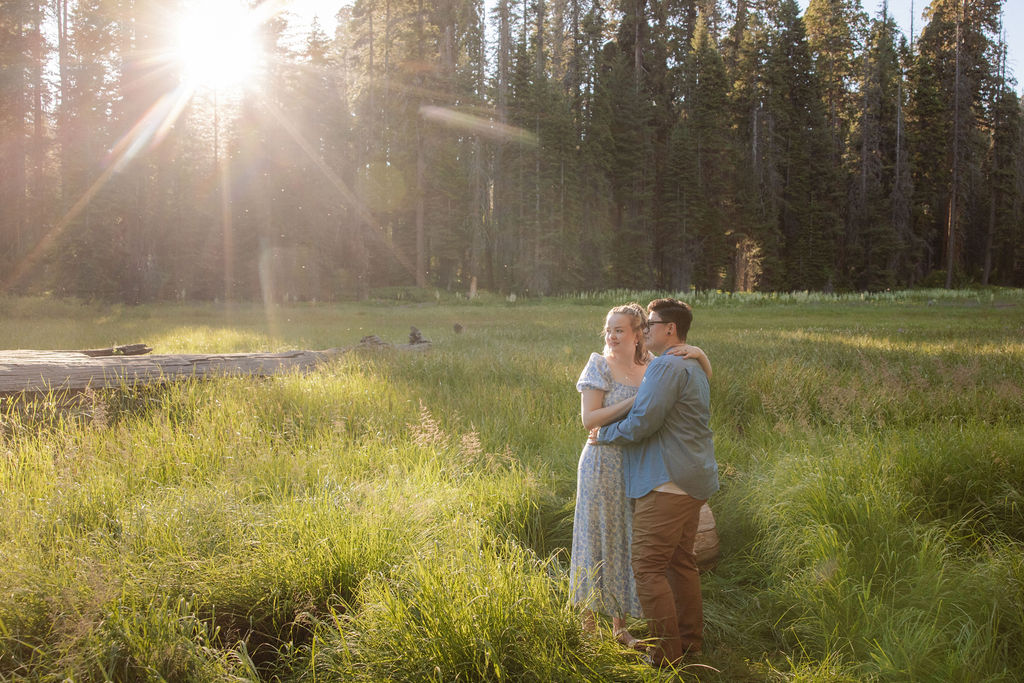 Two people embrace and kiss in a grassy field with sunlight streaming through trees in the background for their Sequoia National Park Engagement Photos