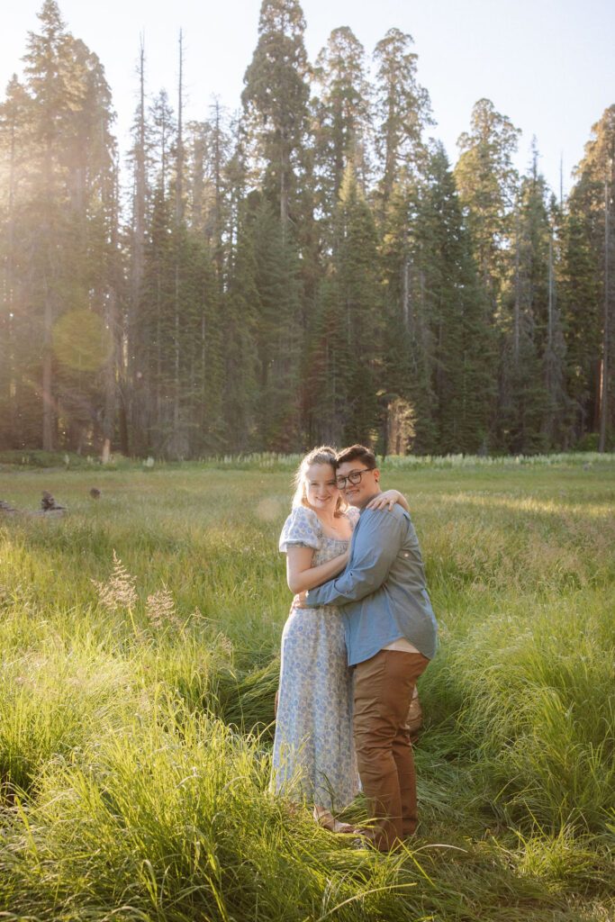 Two people embrace and kiss in a grassy field with sunlight streaming through trees in the background for their Sequoia National Park Engagement Photos