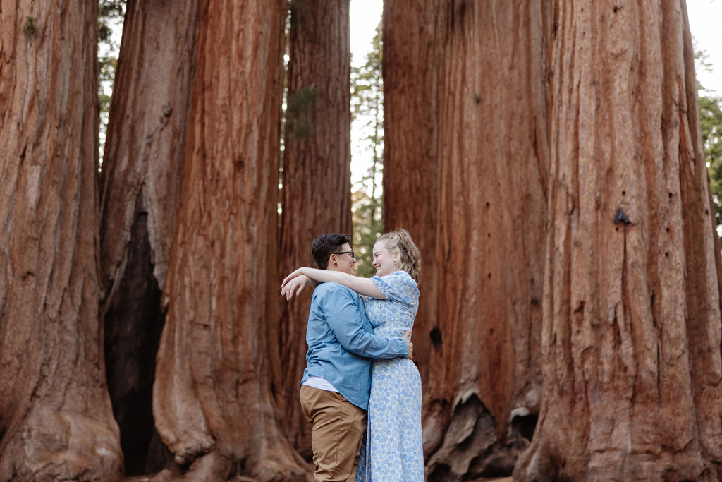 A couple embraces in front of tall redwood trees in an outdoor forest setting.