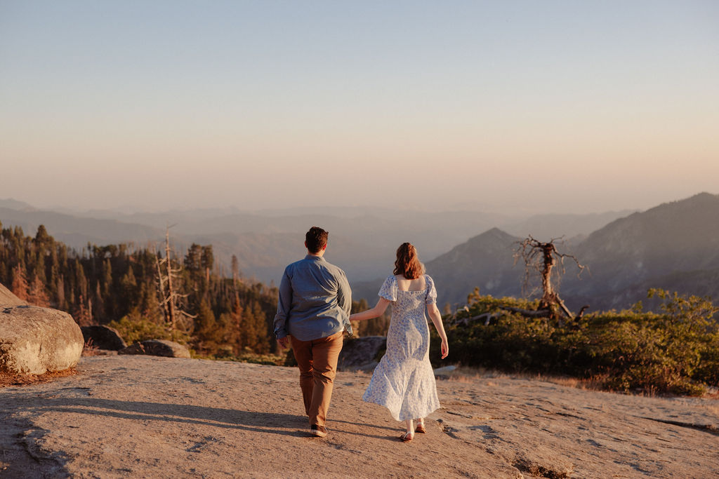A couple holding hands walks on a rocky surface overlooking a scenic mountain landscape at sunset for their sequoia national park engagement photos